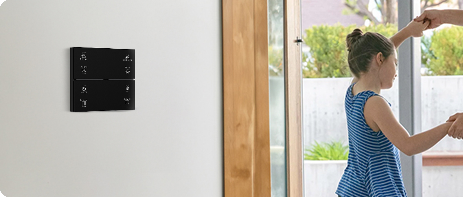 A Pert Stella matte-black glass wall keypad with engraved, backlit Morning, Evening, Good Night, Relax, Master, AC and Curtains buttons beside an open glass door, where a girl dances with her mother in a bright family home
